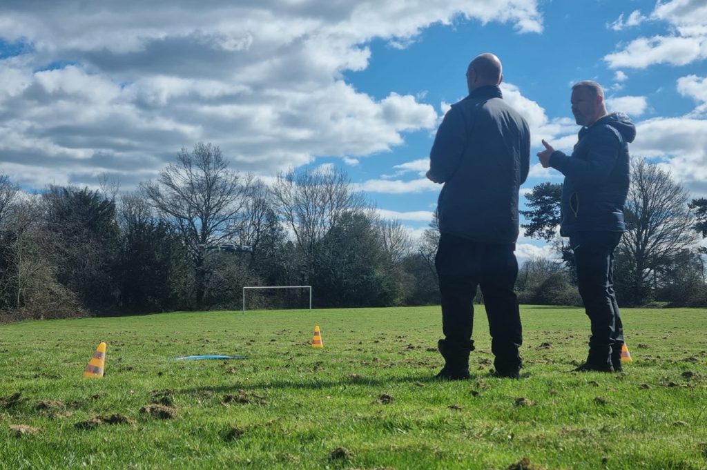 Two men stood outside looking at a drone landing pad. One man is speaking to the other.