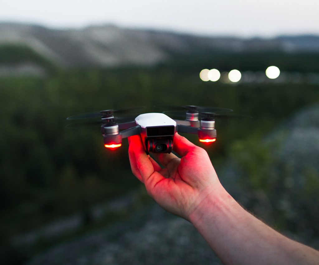 Hand holding a DJI Mini drone with red lights and mountains in the background.